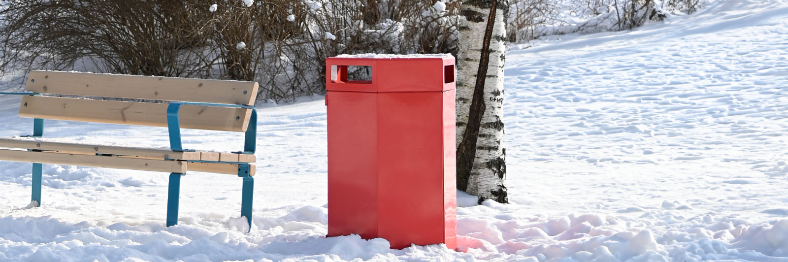 Large red recycling bin situated next to a park bench. It is snowing in the surroundings.